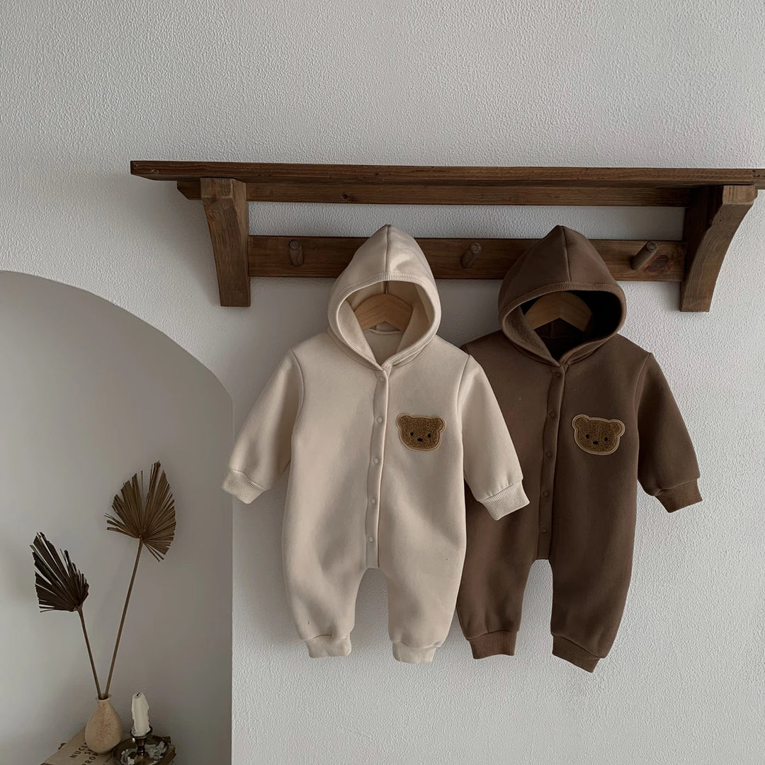 Deux combinaisons bébé beige et marron avec capuche et patch ourson, suspendues sur une étagère en bois.
