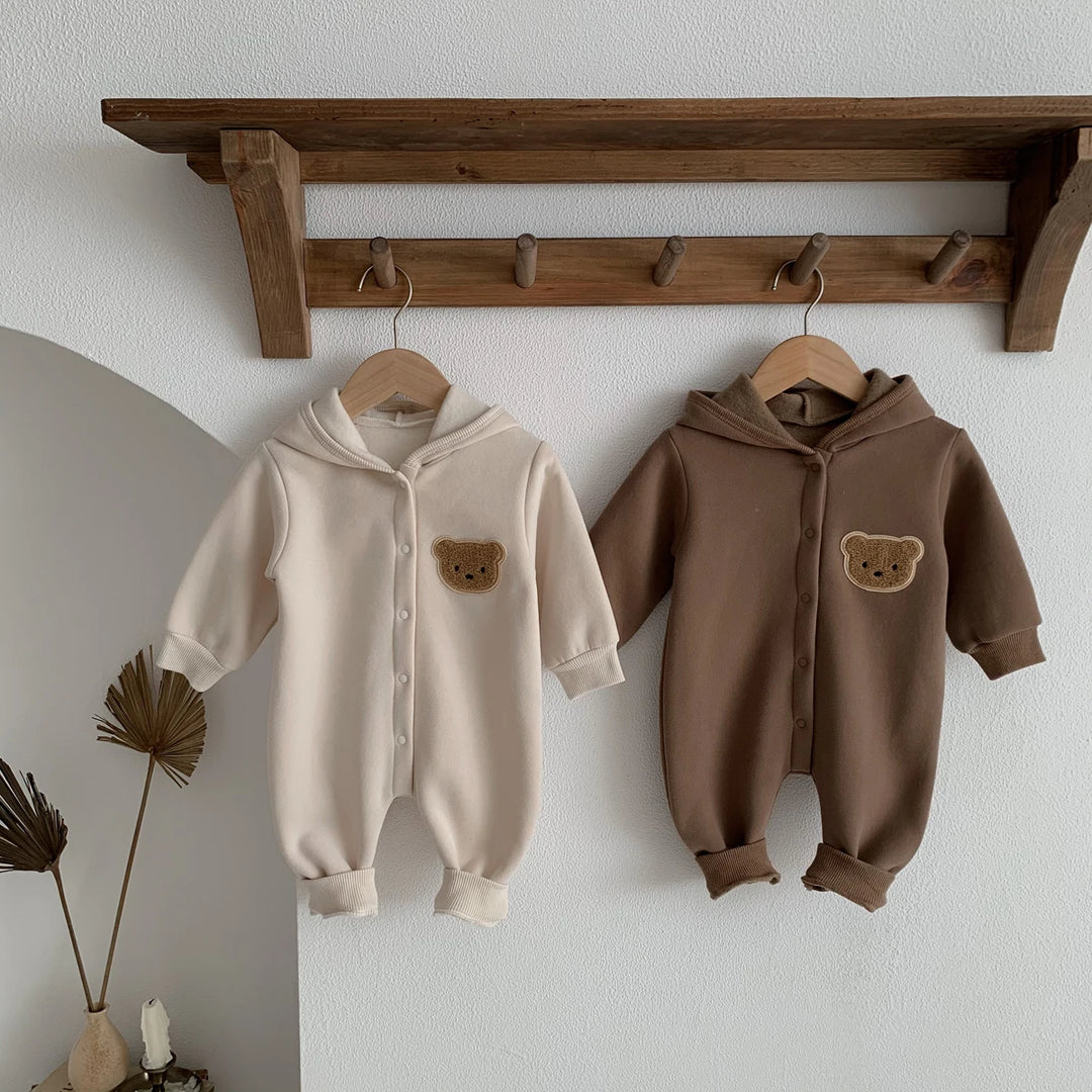 Deux combinaisons bébé beige et marron avec capuche et ourson, suspendues à une étagère décorative.