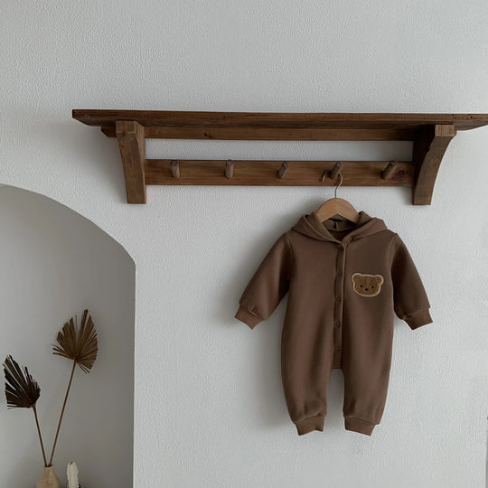 Combinaison bébé marron à capuche avec motif ourson, suspendue sur un cintre sous une étagère en bois.