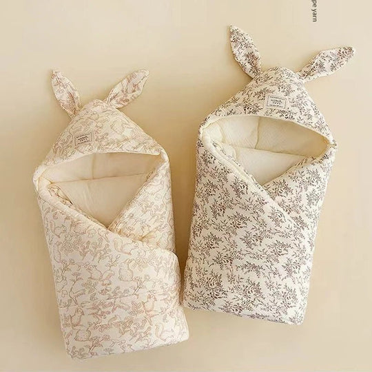 Deux couvertures d’emmaillotage pour bébé en mousseline de coton, couleur ivoire avec motif floral et capuche à oreilles, présentées à plat dans un décor doux et naturel Kalinatur.