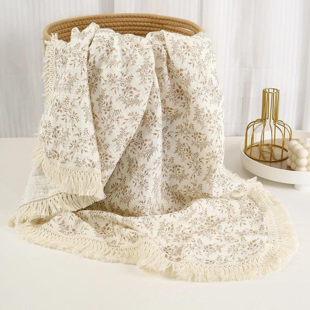 Couverture légère pour bébé Kalinatur, tissu blanc crème à motif feuillage beige avec franges, présentée sur un panier naturel.
