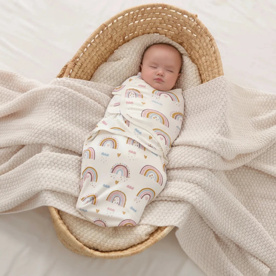 Bébé emmailloté dans une gigoteuse Kalinatur motif arc-en-ciel, couché dans un panier en osier sur une couverture beige.