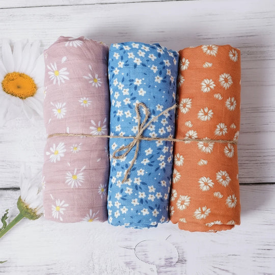 Trois langes en mousseline pour bébé, roulés et attachés, avec motifs floraux violets, bleus et orange, présentés sur un fond en bois blanc.
