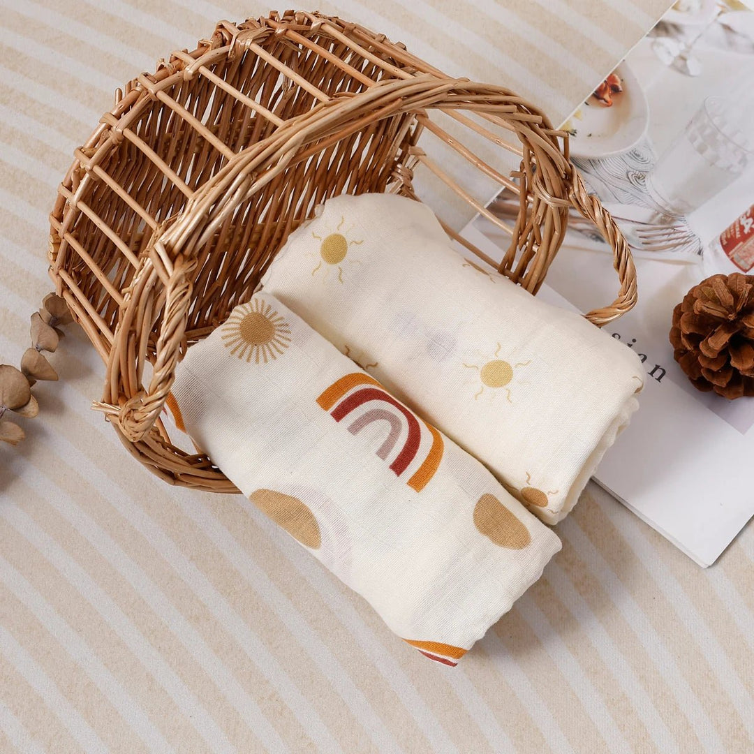 Deux langes en mousseline pour bébé, roulés et présentés dans un panier en rotin, décorés de motifs arc-en-ciel et soleil dans des tons doux.