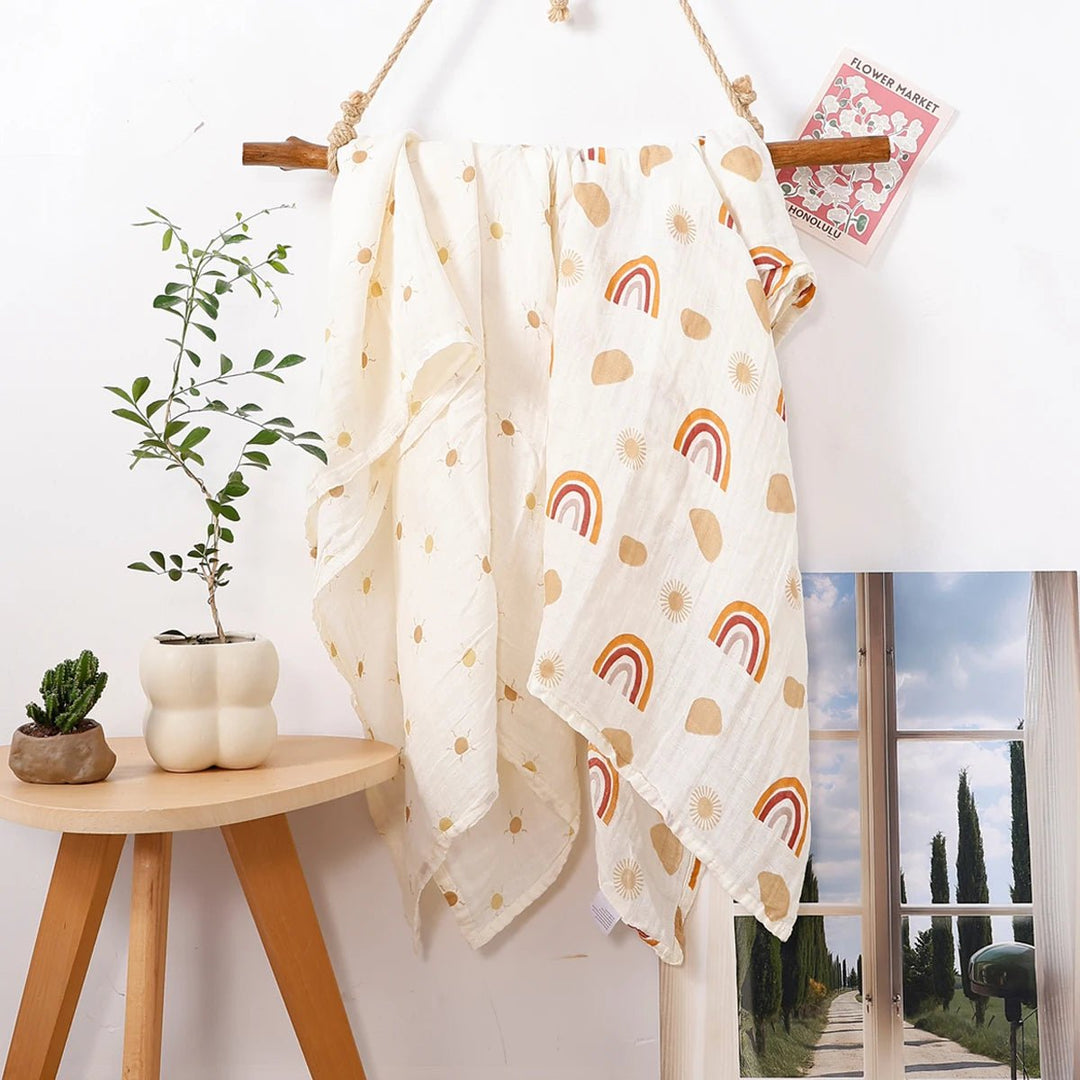 Lange en mousseline pour bébé avec motifs arc-en-ciel et soleil, suspendu sur un support en bois dans un décor naturel.