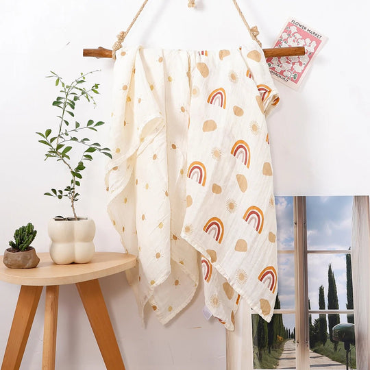 Lange en mousseline pour bébé avec motifs arc-en-ciel et soleil, suspendu sur un support en bois dans un décor naturel.