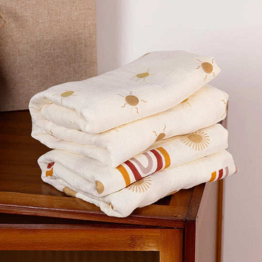 Langes en mousseline pour bébé, pliés et empilés, décorés de motifs soleil et arc-en-ciel dans des tons naturels, posés sur un meuble en bois.