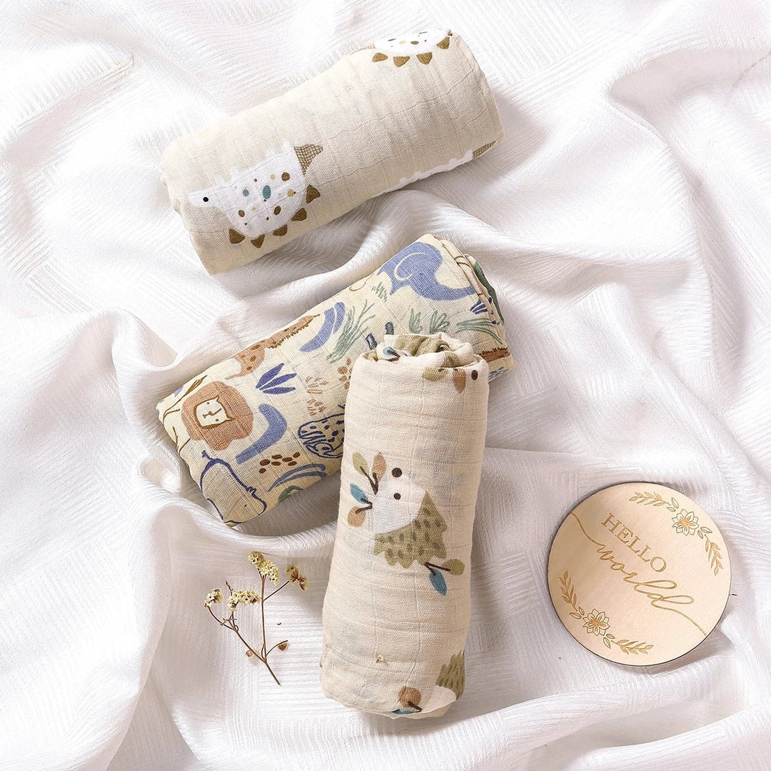 Langes en mousseline pour bébé, roulés et posés sur un drap blanc, décorés de motifs animaux et naturels dans des tons doux, accompagnés d’un disque “Hello world”.