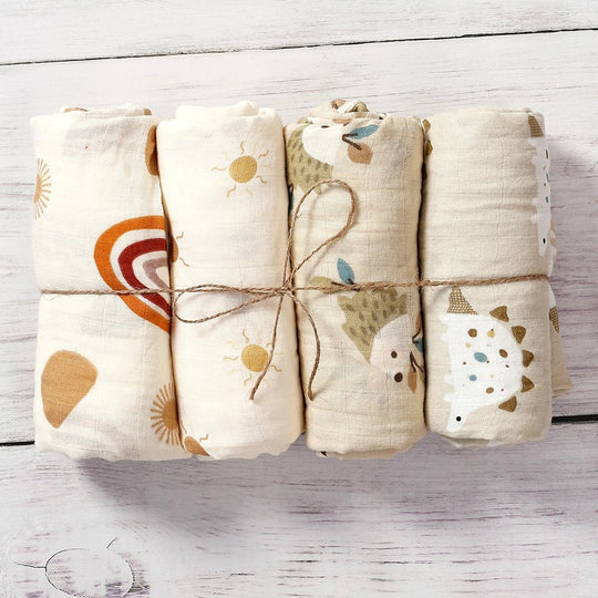 Quatre langes en mousseline pour bébé, roulés et attachés, avec motifs bohèmes et animaux dans des tons naturels, présentés sur un fond en bois blanc.