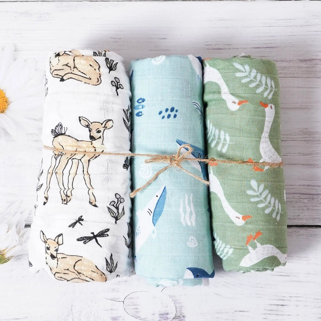 Trois langes en mousseline pour bébé, roulés et attachés, décorés de motifs cerf, plumes et oies dans des tons naturels, présentés sur un fond en bois blanc.