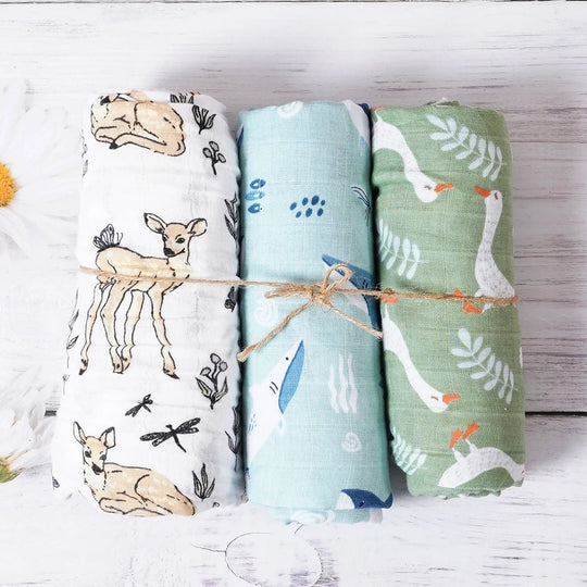 Trois langes en mousseline pour bébé, roulés et attachés, décorés de motifs cerf, plumes et oies dans des tons naturels, présentés sur un fond en bois blanc.