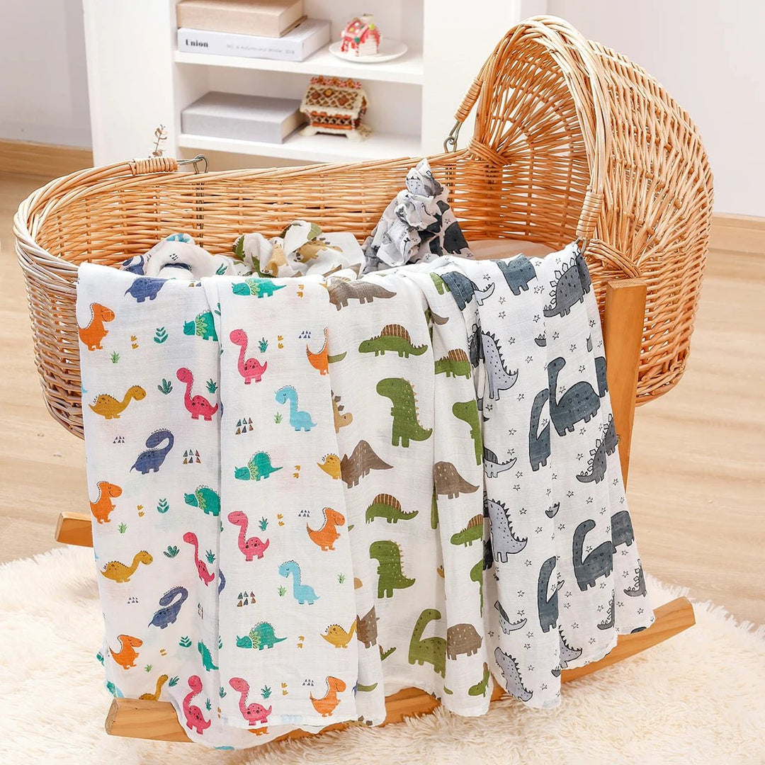 Langes en mousseline pour bébé décorés de motifs dinosaures multicolores, suspendus sur un berceau en rotin dans un décor naturel.