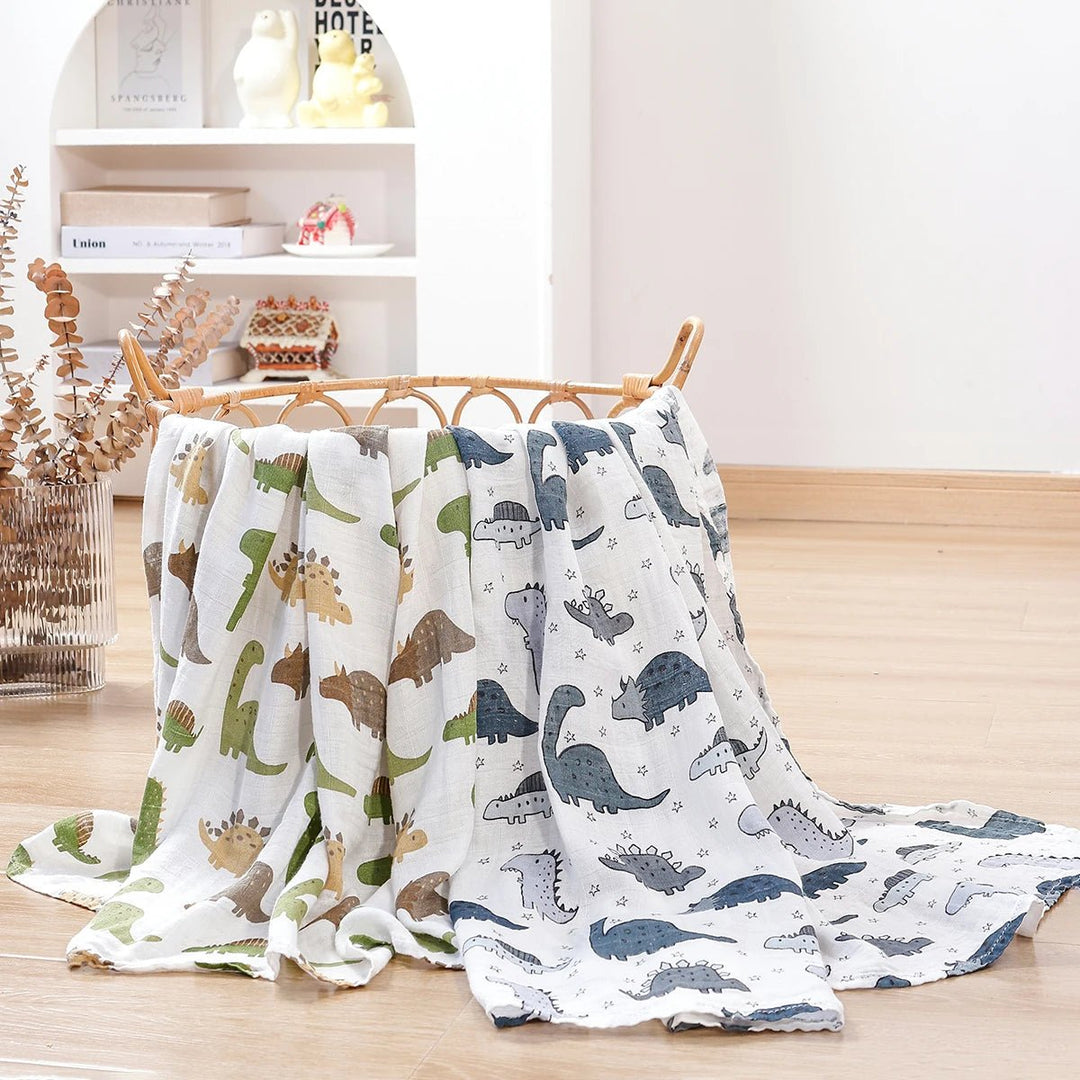 Langes en mousseline pour bébé décorés de motifs dinosaures verts et gris, suspendus sur un panier en rotin dans un décor lumineux.