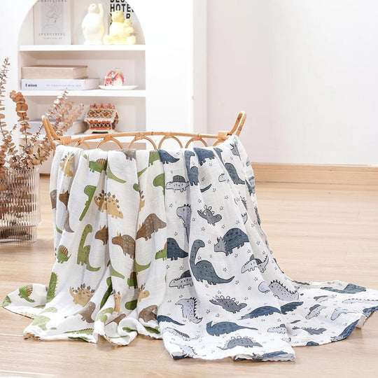 Langes en mousseline pour bébé décorés de motifs dinosaures verts et gris, suspendus sur un panier en rotin dans un décor lumineux.