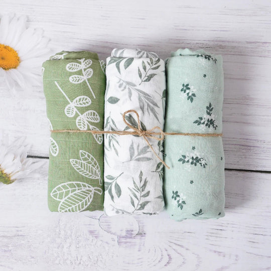 Trois langes en mousseline pour bébé, roulés et attachés, avec motifs feuilles dans des tons verts naturels, présentés sur un fond en bois blanc.