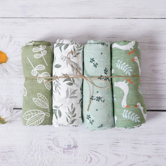 Trois langes en mousseline pour bébé, roulés et attachés, avec motifs feuilles dans des tons verts naturels, présentés sur un fond en bois blanc.