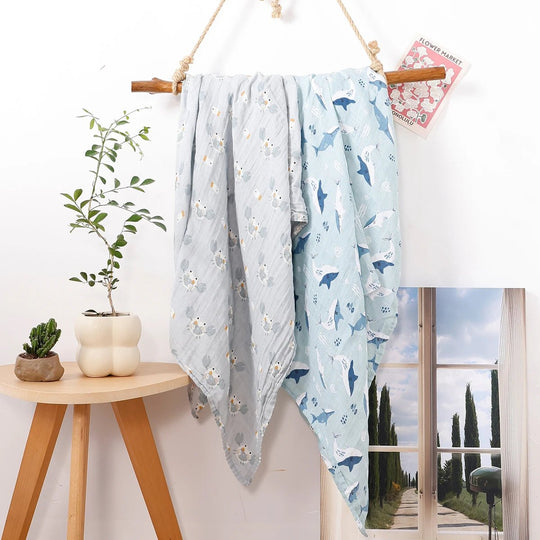 Langes en mousseline pour bébé à motifs marins et dauphins, suspendus sur un support en bois dans un décor naturel et lumineux.