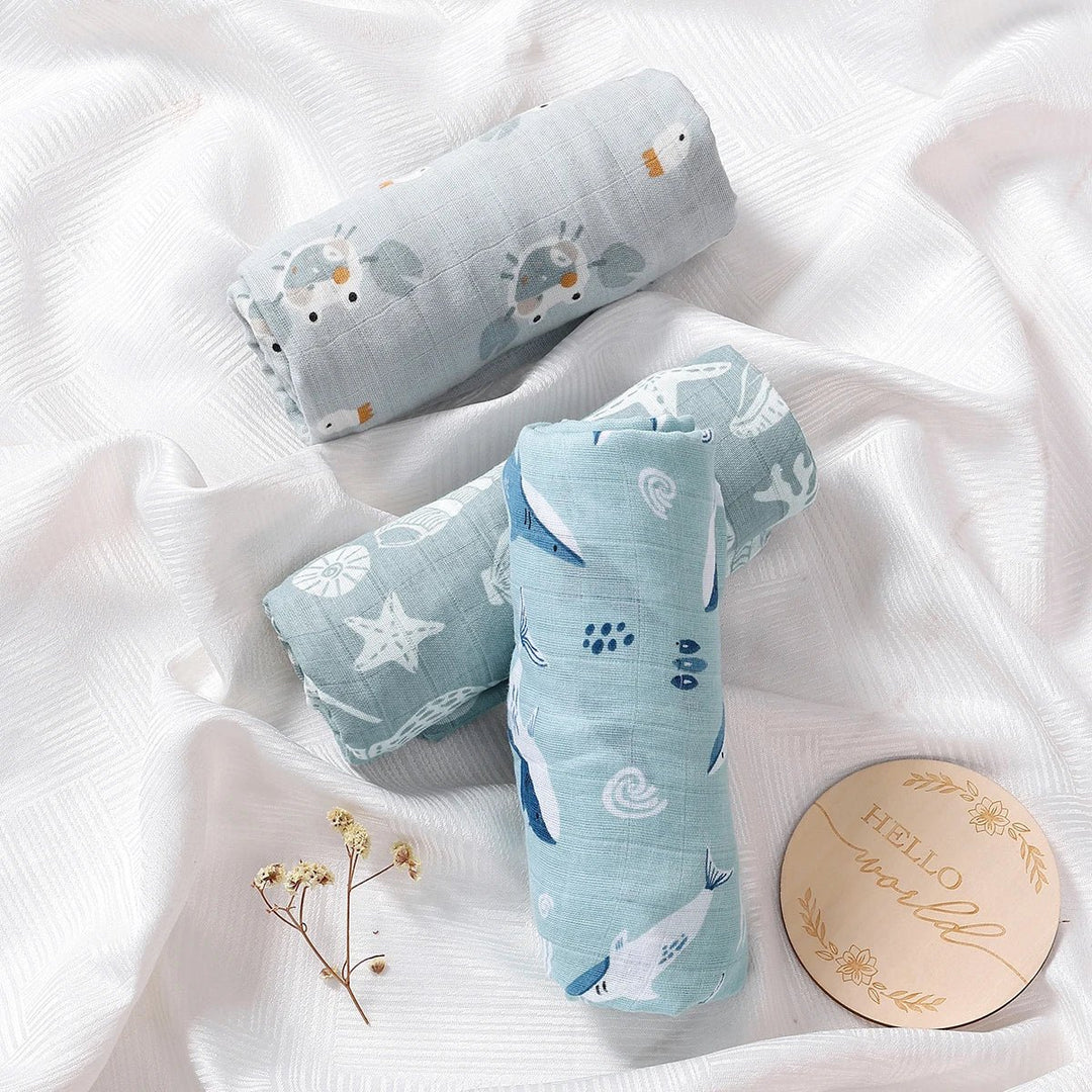 Langes en mousseline pour bébé, roulés et posés sur un drap blanc, décorés de motifs marins avec poissons, étoiles et coquillages, accompagnés d’un disque “Hello world”.