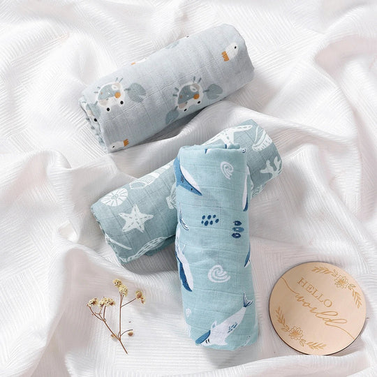 Langes en mousseline pour bébé, roulés et posés sur un drap blanc, décorés de motifs marins avec poissons, étoiles et coquillages, accompagnés d’un disque “Hello world”.