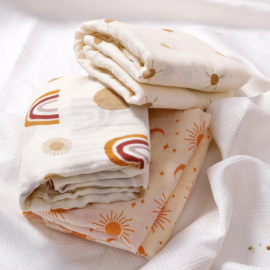 Langes en mousseline pour bébé, pliés et posés sur un tissu blanc, décorés de motifs soleil, lune et arc-en-ciel dans des tons naturels.