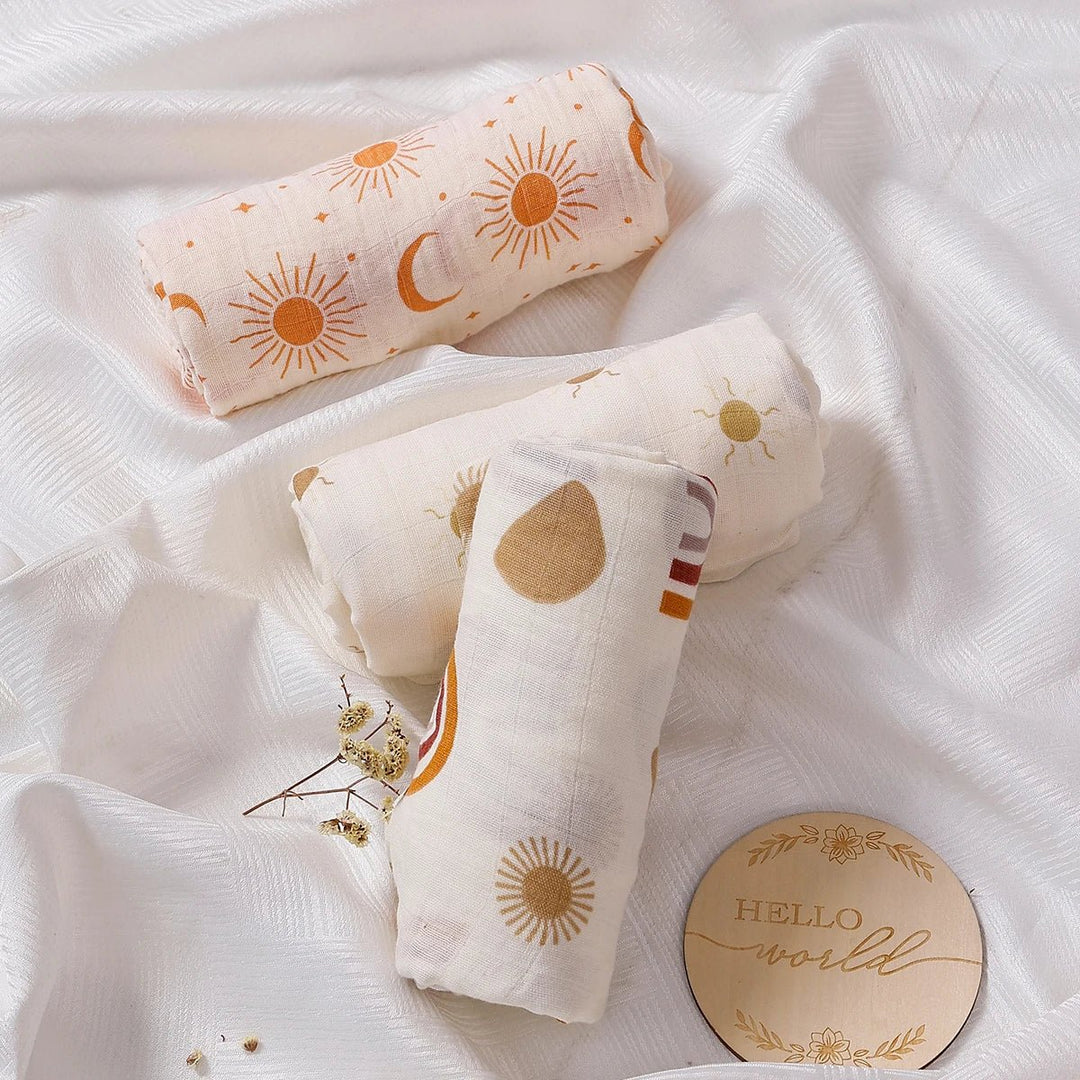 Langes en mousseline pour bébé, roulés et posés sur un tissu blanc, décorés de motifs soleil et lune, accompagnés d’un disque “Hello world”.