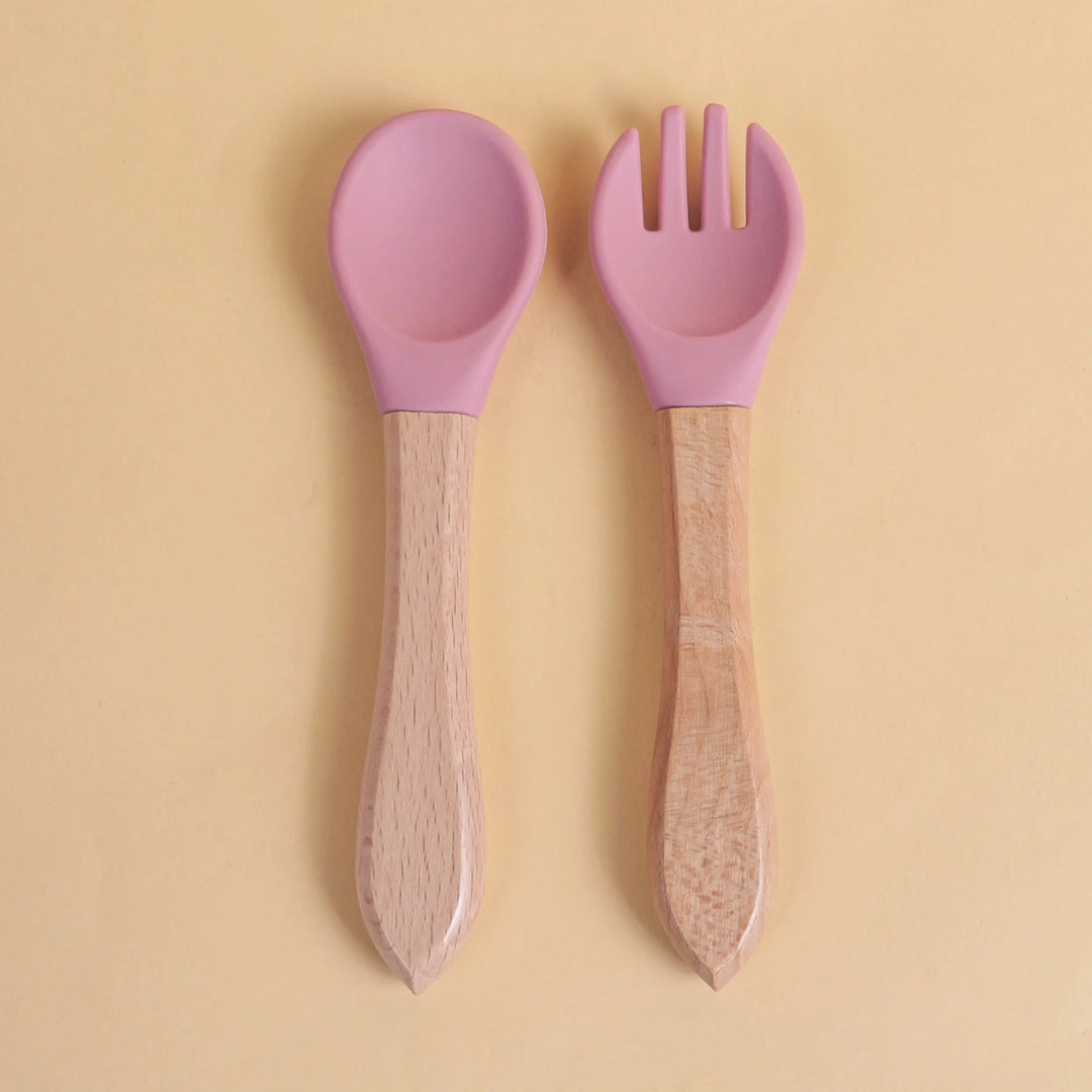 Set de cuillère et fourchette pour bébé en silicone rose avec manche en bois naturel sur fond beige
