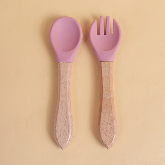 Set de cuillère et fourchette pour bébé en silicone rose avec manche en bois naturel sur fond beige
