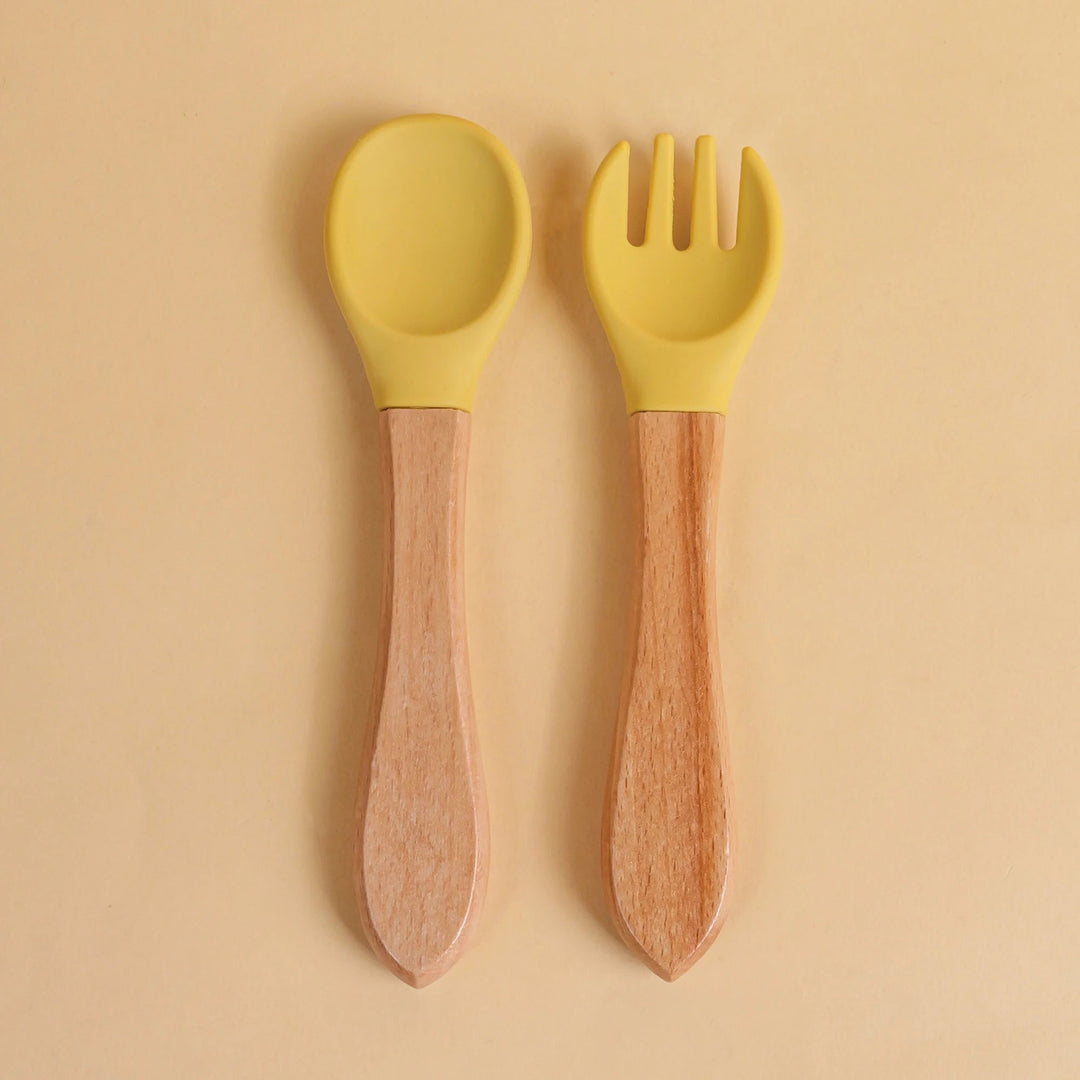 Set de cuillère et fourchette pour bébé en silicone jaune avec manche en bois naturel sur fond beige
