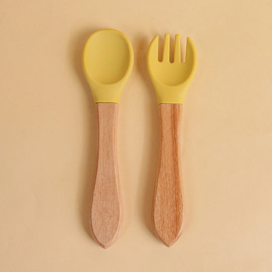 Set de cuillère et fourchette pour bébé en silicone jaune avec manche en bois naturel sur fond beige
