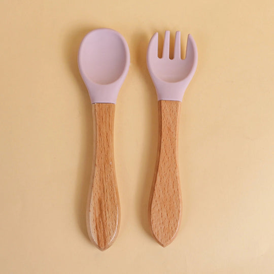 Set de cuillère et fourchette pour bébé en silicone violet avec manche en bois naturel sur fond beige
