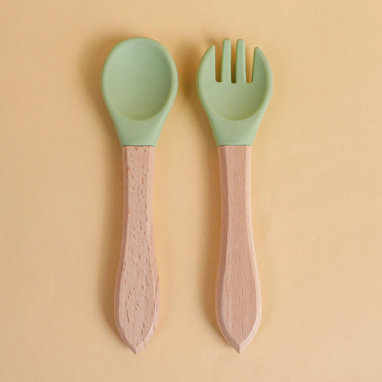Set de cuillère et fourchette pour bébé en silicone vert avec manche en bois naturel sur fond beige

