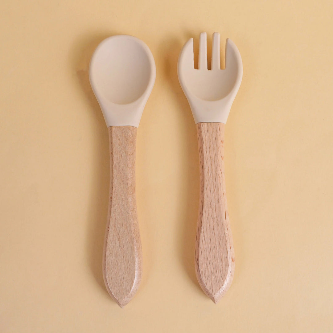 Set de cuillère et fourchette pour bébé en silicone blanc avec manche en bois naturel sur fond beige
