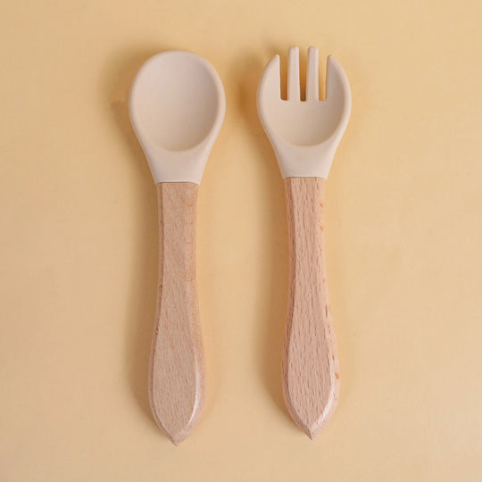 Set de cuillère et fourchette pour bébé en silicone blanc avec manche en bois naturel sur fond beige
